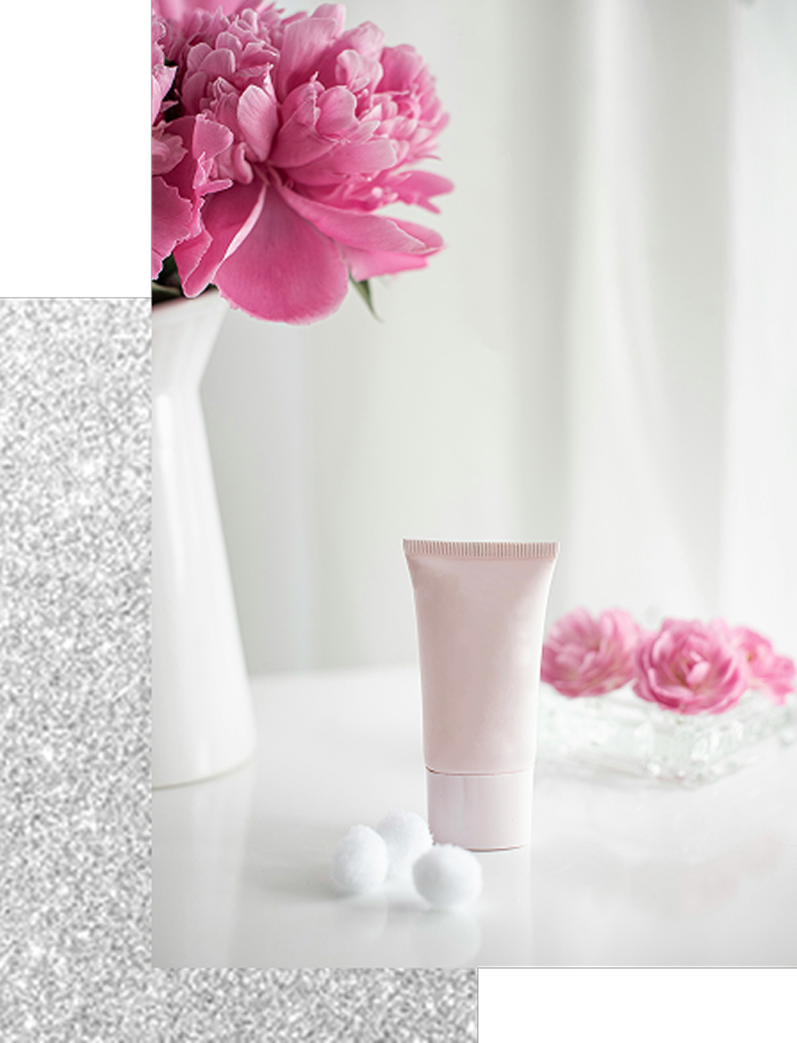 白いテーブルの上に、淡いピンク色のチューブ型コスメとコットンが置かれ、背景にピンクの花が飾られた静物写真